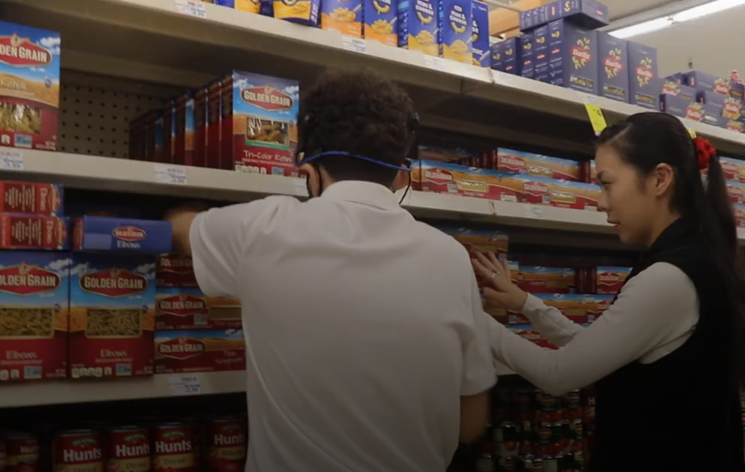 Store Shelf Worker Two individuals are actively stocking shelves in what appears to be a grocery store or supermarket. A person in a white shirt and blue face mask is reaching for items on a lower shelf on the left, while a person with long dark hair, wearing a dark vest over a light long-sleeved top, is also placing items on a shelf on the right. The shelves are filled with boxes of pasta (specifically 'GOLDEN GRAIN' brand) and cans of 'Hunts' tomatoes.