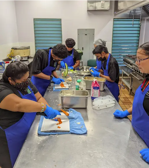 A group of young individuals, wearing black shirts and blue aprons with blue gloves, are actively engaged in food preparation on a stainless steel counter in a commercial kitchen setting. They appear to be chopping ingredients and using various kitchen tools.