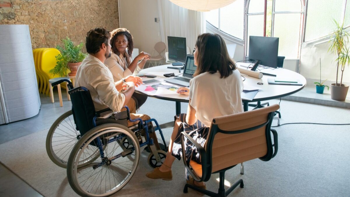 An inclusive and modern office that has a person in a wheelchair and two people sitting beside him having a meeting.